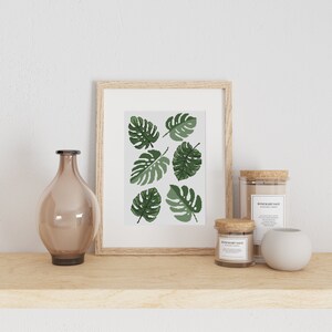 Op de afbeelding: Een ingelijst print met een witte achtergrond en een patroon van groene monstera bladeren. De print staat op een houten plank met een glazen vaas, twee kaarsen en een kleine witte keramische container.
