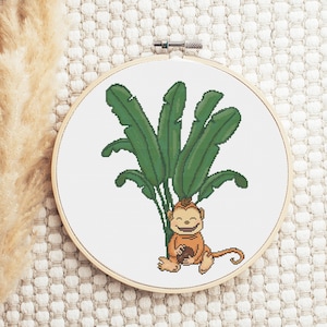 Op de afbeelding: Een borduurring met kruissteek met een glimlachende aap die onder een bananenboom zit. De aap houdt een bruin voorwerp in zijn handen. De bananenboom is groen tegen een witte achtergrond.