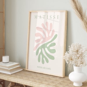 Puede incluir: Una impresión enmarcada con un diseño abstracto en verde y rosa. La impresión está titulada "Matisse" y "Papiers Découpés". La impresión está colgada en una pared verde claro sobre un banco de madera con una parte superior de ratán tejido.