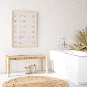May include: A white bathtub with a chrome faucet and a palm leaf plant in the background. A wooden bench with a woven seat sits in front of a framed print with a pattern of abstract symbols.