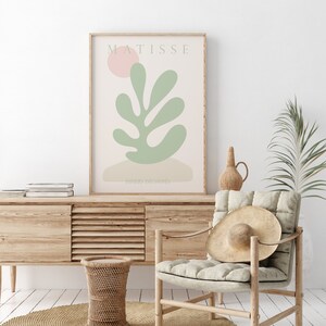 Op de afbeelding: Een ingelijst afdruk van een Matisse-kunstwerk met een groen blad en een roze cirkel op de achtergrond. De tekst "MATISSE" en "PAPIERS DECOUPES" is zichtbaar op de afdruk.