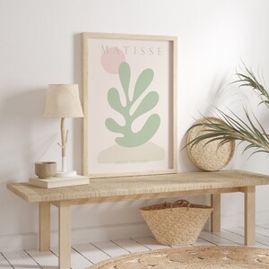 Op de afbeelding: Een ingelijste print van een Matisse-kunstwerk met een groen blad met een roze cirkel op de achtergrond. De print is getiteld "Papiers Decoupes" en wordt tentoongesteld op een houten bank met een geweven tapijt eronder.