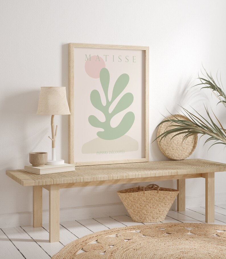 Op de afbeelding: Een ingelijste print van een Matisse-kunstwerk met een groen blad met een roze cirkel op de achtergrond. De print is getiteld "Papiers Decoupes" en wordt tentoongesteld op een houten bank met een geweven tapijt eronder.