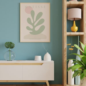 Op de afbeelding: Een wit dressoir met een houten blad en poten, een ingelijst afdruk van een Matisse-schilderij met de tekst "MATISSE" en "PAPIERS DECOUPES", een vaas met een groene plant, een witte vaas met een droge tak, een wit keramiek object, een houten plank met een roze lamp met een gele voet, een vaas met gele bloemen, een vaas met een groene plant en een witte vaas.