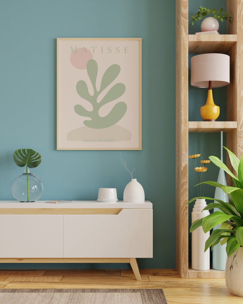 Op de afbeelding: Een wit dressoir met een houten blad en poten, een ingelijst afdruk van een Matisse-schilderij met de tekst "MATISSE" en "PAPIERS DECOUPES", een vaas met een groene plant, een witte vaas met een droge tak, een wit keramiek object, een houten plank met een roze lamp met een gele voet, een vaas met gele bloemen, een vaas met een groene plant en een witte vaas.