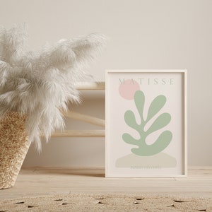 Op de afbeelding: Een witte ingelijste print met een roze achtergrond en een groen bladerenontwerp. De print heeft de tekst "MATISSE" en "PAPIERS DECOUPES" in zwart.