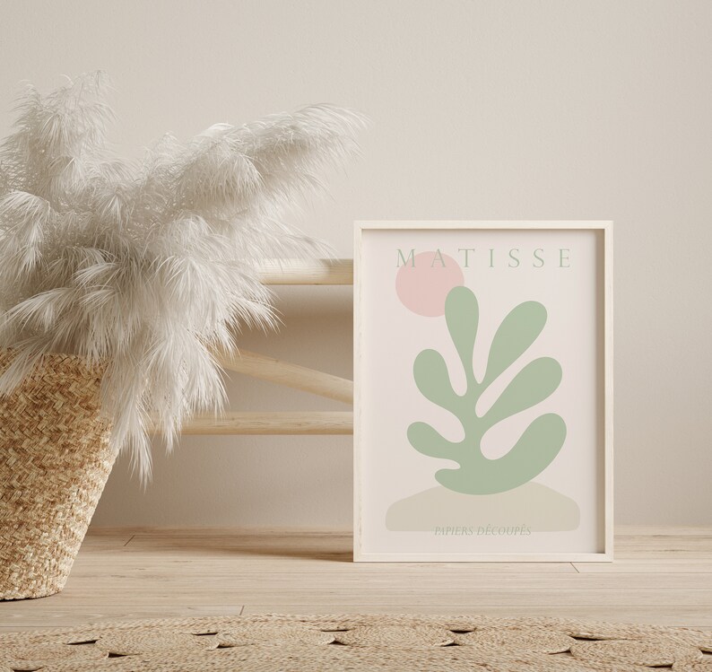 Op de afbeelding: Een witte ingelijste print met een roze achtergrond en een groen bladerenontwerp. De print heeft de tekst "MATISSE" en "PAPIERS DECOUPES" in zwart.