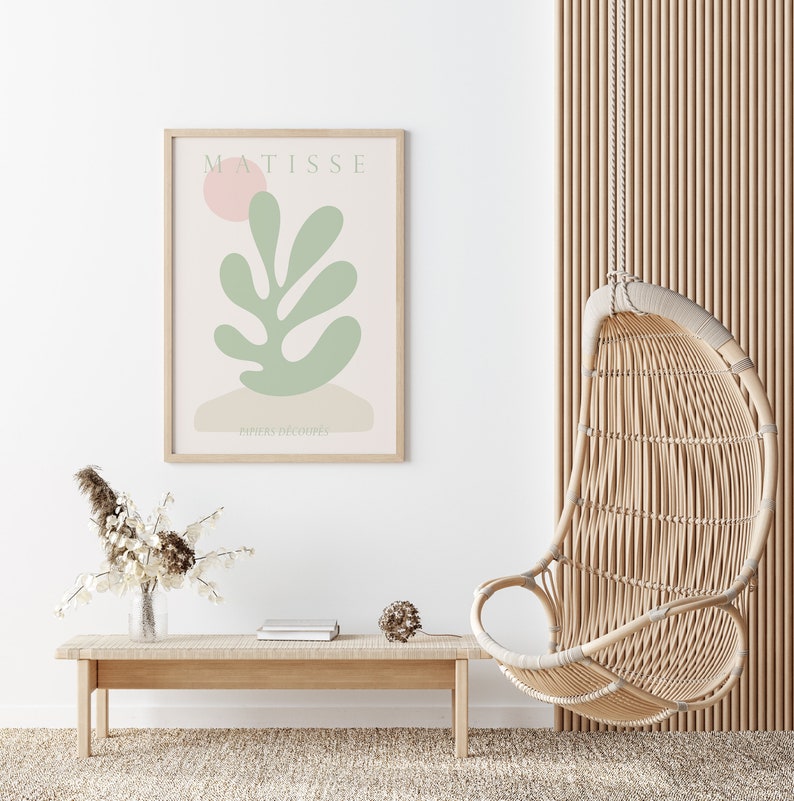 Op de afbeelding: Een lichte houten bank met een boek en een kleine decoratieve bal erop. Op de achtergrond is een hangstoel van gevlochten riet te zien met een witte muur en een ingelijst afdruk van een groen blad met de tekst "MATISSE PAPIERS DECOUPES" erop.