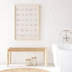 May include: A white bathroom with a white bathtub, a wooden bench with a woven seat, and a framed print with a series of symbols on a white background. Two glass lanterns with rope accents are on the floor in front of the bench.
