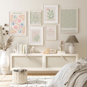 Op de afbeelding: Een witte commode met twee laden en een witte lamp met een grijze kap. Boven de commode hangen vijf ingelijste prints aan de wall. De prints zijn van verschillende bloem- en abstracte ontwerpen. De prints zijn in een minimalistische stijl met een licht kleurenpalet. De commode is versierd met een rieten mand, een schelp en twee witte keramische dierenfiguren.