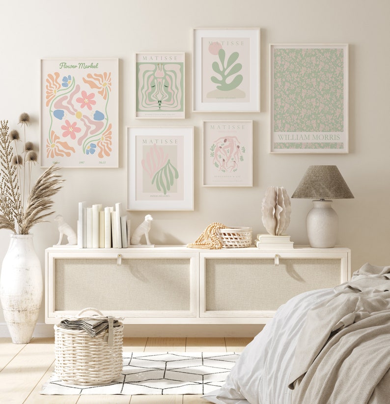 Op de afbeelding: Een witte commode met twee laden en een witte lamp met een grijze kap. Boven de commode hangen vijf ingelijste prints aan de wall. De prints zijn van verschillende bloem- en abstracte ontwerpen. De prints zijn in een minimalistische stijl met een licht kleurenpalet. De commode is versierd met een rieten mand, een schelp en twee witte keramische dierenfiguren.