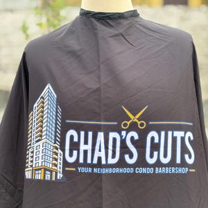 May include: A black barber cape featuring a graphic of a tall skyscraper building, a pair of yellow scissors, and the text CHAD'S CUTS YOUR NEIGHBORHOOD CONDO BARBERSHOP printed in white and yellow on the front of the fabric draped over a mannequin.