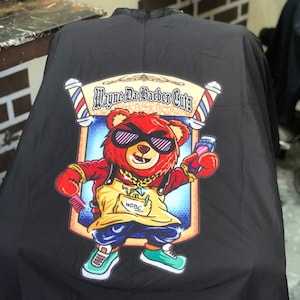 May include: Black barber cape with a cartoon bear wearing sunglasses and holding a razor. The bear is wearing a yellow vest with the text "WDBC" on it. The text "Wayne Da Barber Cutz" is above the bear.