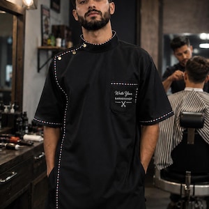 Może przedstawiać: Czarna tunika fryzjerska z krótkim rękawem, wysokim kołnierzem ze złotymi guzikami, kieszenią na piersi z napisem "Write Your BARBERSHOP" i ikoną nożyczek oraz czerwono-biało-niebieską lamówką w paski wzdłuż kołnierza, kieszeni i szwu bocznego.