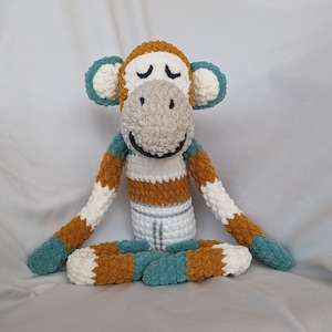 May include: A crocheted monkey toy sitting in a meditative pose with its eyes closed. The monkey is brown, white, and teal with a striped pattern on its torso.