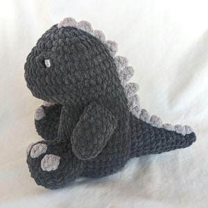 May include: A gray crocheted dinosaur plush toy with a light gray belly and spikes. The dinosaur is sitting on a white surface.