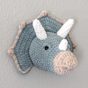 May include: A soft, blue triceratops head with white horns and a beige trim. The dinosaur features a pale pink snout and a black eye. This decorative item is suitable for a child's room or nursery.