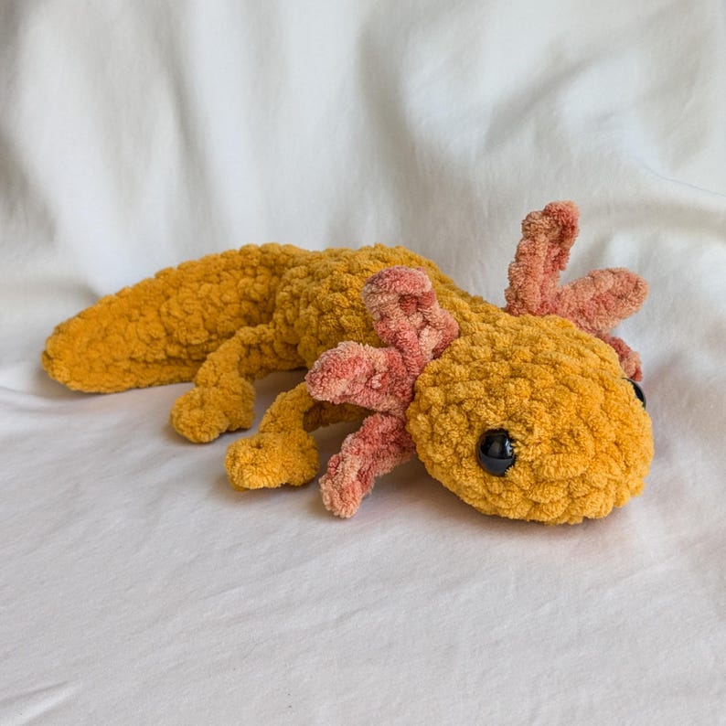 Puede incluir: Un peluche de ajolote hecho a mano en tonos amarillos y rosas. El cuerpo es de un amarillo dorado texturizado, con una cola larga y cuatro patas. Las branquias y los detalles de la cabeza son de un rosa suave. Ojos negros completan el dise&ntilde;o.