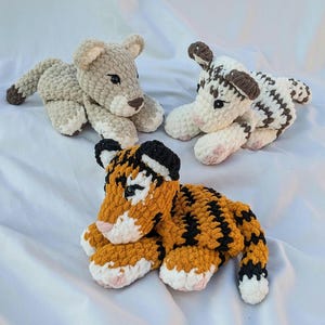 May include: Three crocheted stuffed animals: a brown and white striped tiger, a light brown lion, and a white and brown zebra. The animals are lying on a white surface.