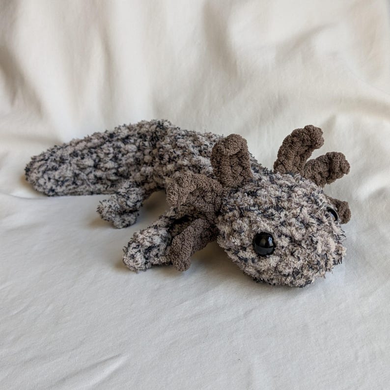 Puede incluir: Un peluche de ajolote hecho a mano en tonos grises y marrones. El peluche tiene un cuerpo texturizado y difuso, ojos oscuros y branquias detalladas. El ajolote est&aacute; tumbado sobre una superficie blanca.