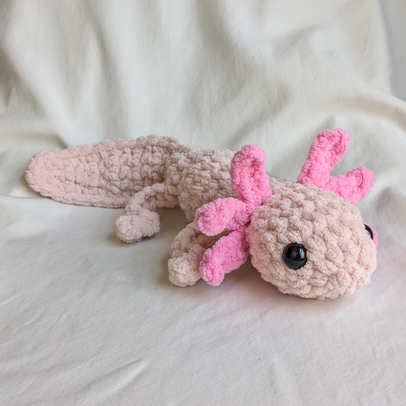 Puede incluir: Un peluche de ajolote hecho a mano en tonos rosa y beige. El ajolote presenta un cuerpo texturizado y difuso, con branquias y acentos de color rosa brillante. Tiene ojos grandes, negros y redondos, y est&aacute; sobre una superficie blanca.
