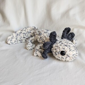 Puede incluir: Un peluche de ajolote en blanco con motas azul marino. El ajolote tiene branquias de color gris oscuro y un cuerpo gris oscuro. El juguete tiene un ojo de bot&oacute;n negro. El peluche est&aacute; sobre una superficie blanca.