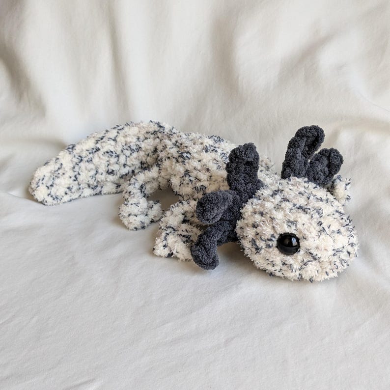 Puede incluir: Un peluche de ajolote en blanco con motas azul marino. El ajolote tiene branquias de color gris oscuro y un cuerpo gris oscuro. El juguete tiene un ojo de bot&oacute;n negro. El peluche est&aacute; sobre una superficie blanca.