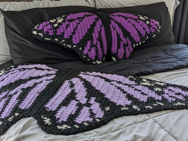 Peut inclure: Un oreiller de jet en forme de papillon crochet&eacute; en violet et noir sur un lit. L'oreiller est en forme de papillon avec ses ailes d&eacute;ploy&eacute;es.