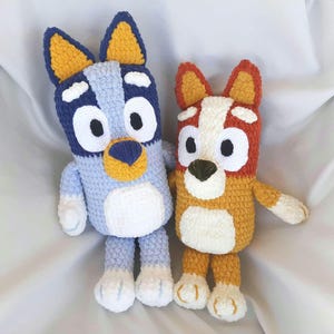 May include: Two handmade crochet plush toys representing cartoon dogs. The larger figure is light blue and dark blue with yellow ears. The smaller figure is orange and white with yellow ears. Both have large white eyes and white belly patches.