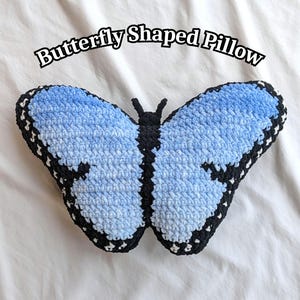 Op de afbeelding: Een gehaakt vlindervormig kussen met lichtblauwe vleugels, zwarte randen en witte accenten. Het centrale lichaam en de voelsprieten zijn zwart. De tekst "Butterfly Shaped Pillow" staat in zwarte letters boven het object.