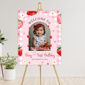 May include: A pink and white welcome sign for a first birthday party. The sign features a photo of a baby girl, strawberries, and daisies. Text includes "Welcome to", "Alexandra's", "Berry First Birthday", and the date November 25, 2025.