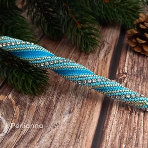 May include: A beaded pen with a turquoise and silver design. The pen is decorated with small beads and rhinestones, with a gold-colored tip. The pen is lying on a wooden surface with pine needles and a pine cone in the background.