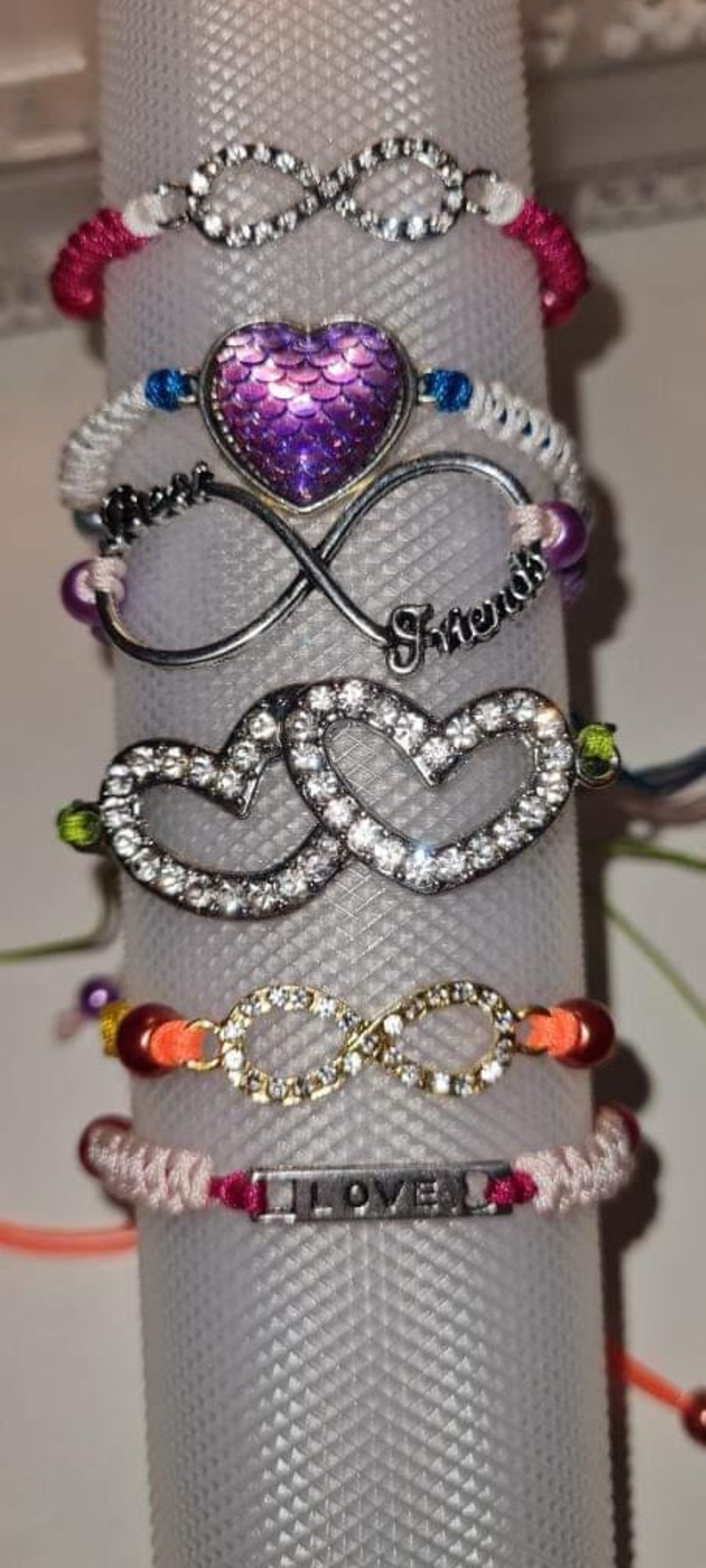 May include: A stack of friendship bracelets with various charms, including infinity symbols, hearts, and a purple mermaid scale charm. The bracelets are made with colourful string and beads.