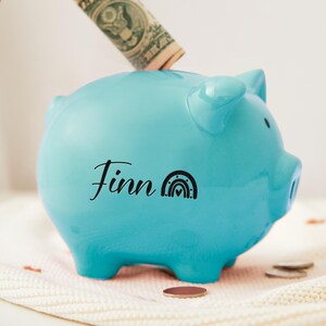 Puede incluir: Una alcancía azul claro con el nombre "Finn" y un gráfico de arcoíris. Una mano está insertando un billete en la ranura superior. La alcancía está sobre una superficie blanca con monedas.