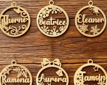 May include: Six wooden Christmas ornaments with names: Thorne, Beatrice, Eleanor, Ramona, Aurora, and Jamsin. Each ornament is circular with a decorative cutout design and a small hole for hanging. The names are laser-cut in a script font.