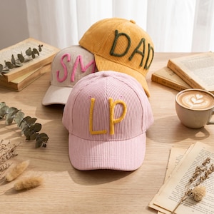 May include: Three corduroy baseball caps in light pink, beige, and mustard yellow. The front-most pink cap features the yellow embroidered letters LP. The beige cap has pink SM letters, and the mustard yellow cap has green DAD letters.