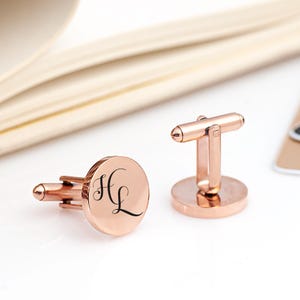 May include: A pair of rose gold cufflinks with a circular design. The cufflinks are engraved with the initials "HL" in a cursive font.