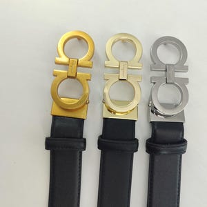 May include: Three black leather belts featuring Gancini-style metal buckles. The left buckle is matte gold, the centre buckle is polished gold, and the right buckle is polished silver. Each buckle displays the Ferragamo brand name engraved on the bar.