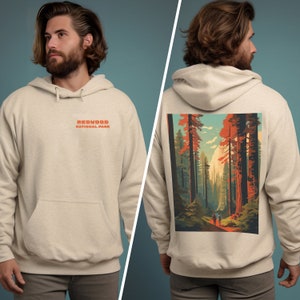 May include: Beige hoodie with a front pocket and the text "REDWOOD NATIONAL PARK" in orange. The back features a colorful illustration of a redwood forest with two people walking on a path. The hoodie is made of a soft material.