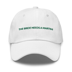 Puede incluir: Gorra de béisbol blanca con el texto "THE BRIDE NEEDS A MARTINI" bordado en verde.