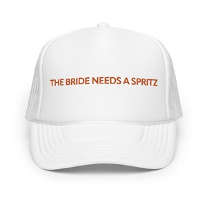 Puede incluir: Gorra de camionero blanca con una frase bordada en rojo "THE BRIDE NEEDS A SPRITZ" en la parte delantera.