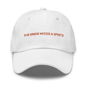 Puede incluir: Gorra de béisbol blanca con el texto "THE BRIDE NEEDS A SPRITZ" bordado en naranja.