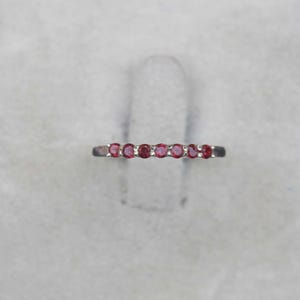 May include: A silver ring featuring a row of seven small, round, bright red gemstones. The ring is set against a soft, neutral background, highlighting the vibrant color of the stones. The ring is a delicate piece of jewelry.