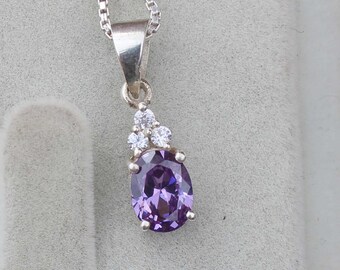 Puede incluir: Un collar con colgante de plata con una gran piedra preciosa ovalada púrpura. El colgante está adornado con tres pequeñas piedras preciosas transparentes. El collar tiene una fina cadena de plata.