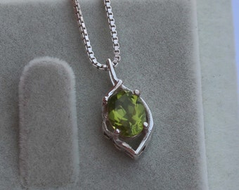 May include: A silver pendant necklace featuring a pear-shaped, light green peridot gemstone. The gemstone is set in a decorative silver frame and hangs from a delicate silver chain. The necklace is displayed on a gray surface.
