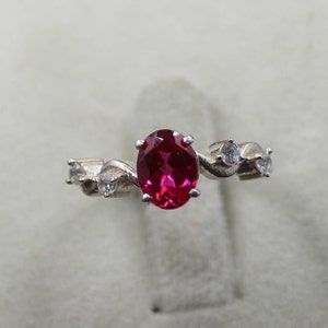 Puede incluir: Un anillo de plata con una gran piedra roja ovalada y piedras más pequeñas transparentes en la banda.