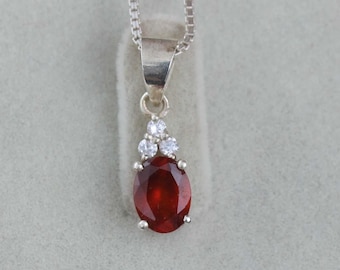 May include: A silver pendant necklace featuring a large, oval-shaped, deep red gemstone. Three smaller, clear gemstones are set above the main stone. The necklace chain is a delicate silver link.