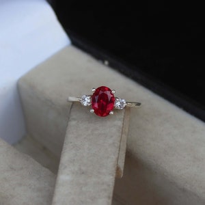 Puede incluir: Anillo de plata con una gema central de rubí ovalada de color rojo intenso, flanqueada por dos gemas claras redondas más pequeñas. El rubí es el punto focal, sobre un fondo neutro.
