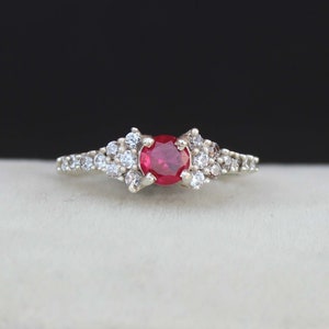May include: A silver ring with a red gemstone surrounded by smaller white gemstones.