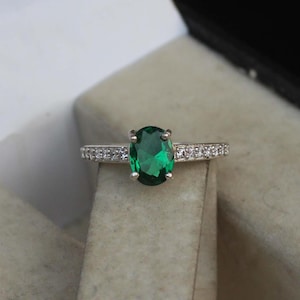 Può includere: Un anello d'argento con una grande pietra preziosa verde di forma ovale circondata da piccole pietre preziose chiare.